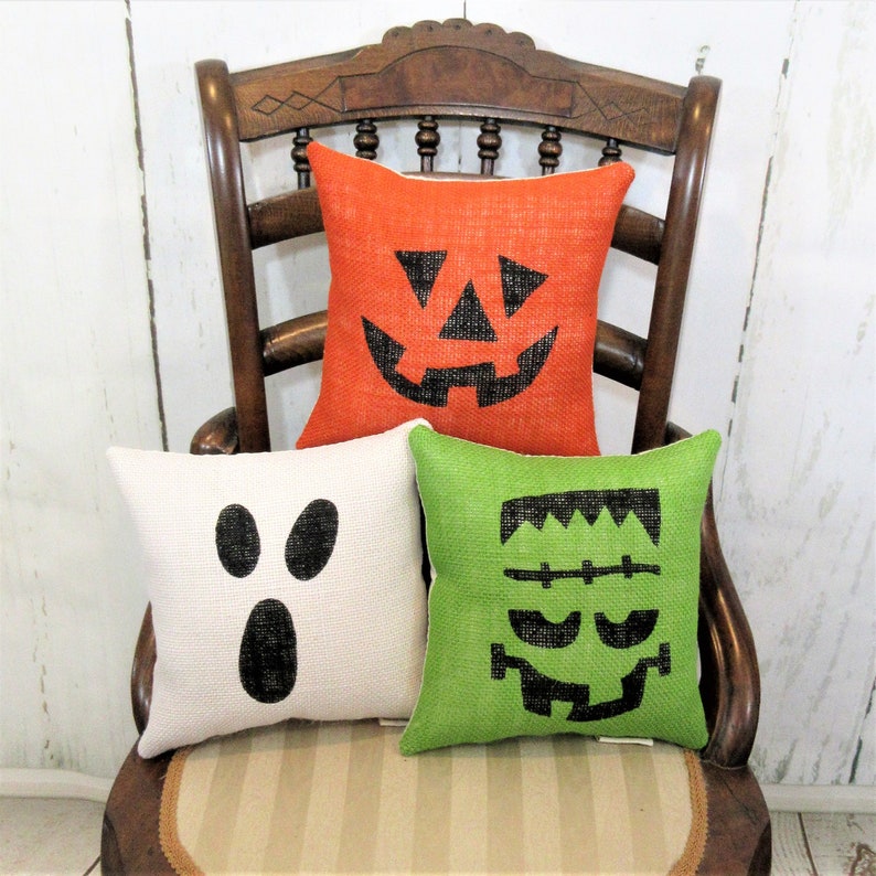 May include: Three Halloween-themed throw pillows with burlap fabric. The pillows feature a pumpkin, ghost, and Frankenstein face in black on orange, white, and green fabric.
