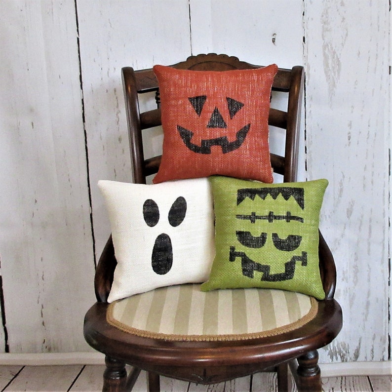May include: Three Halloween-themed throw pillows with burlap fabric and black and white face designs. The pillows are arranged on a wooden chair with a striped cushion.