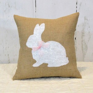 Puede incluir: Una almohada de arpillera con una silueta de conejo blanco y un lazo rosa. La almohada es perfecta para la decoraci&oacute;n de Pascua.