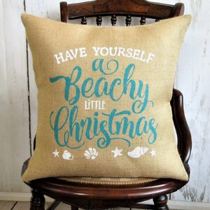 May include: A burlap pillow with the text "Have Yourself a Beachy Little Christmas" in teal blue lettering. The pillow has white starfish, seashells, and a seashell with a white ribbon on it.