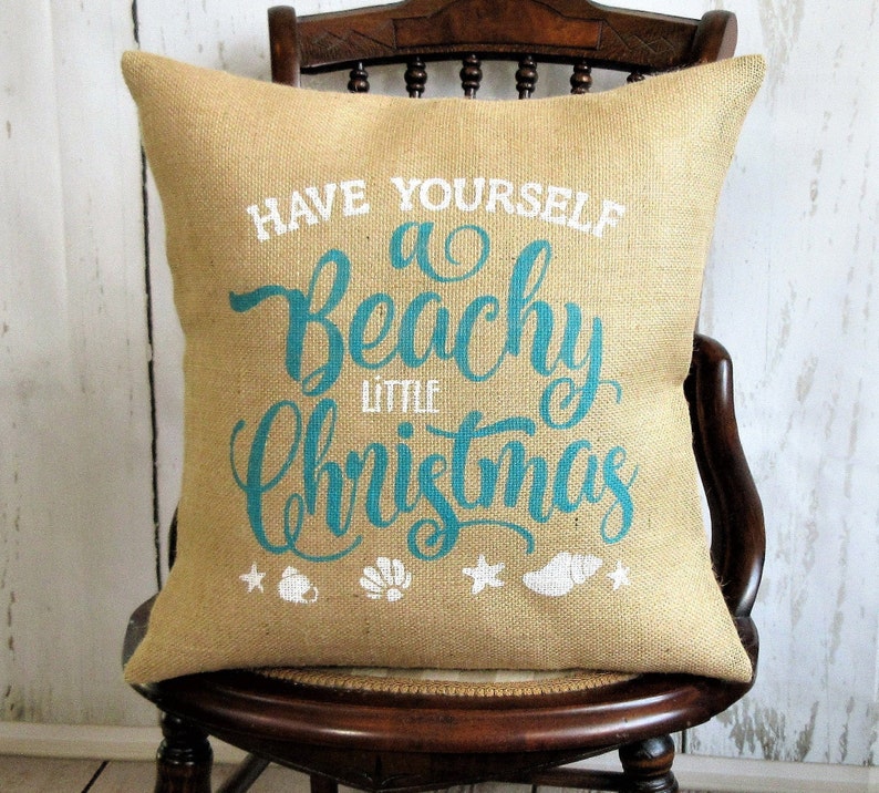 May include: A burlap pillow with the text "Have Yourself a Beachy Little Christmas" in teal blue lettering. The pillow has white starfish, seashells, and a seashell with a white ribbon on it.