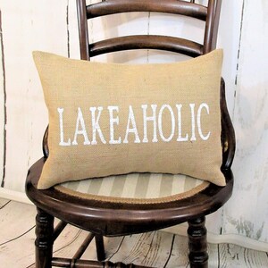 May include: A brown burlap pillow with white text that reads "LAKEAHOLIC" on a wooden chair with a striped seat.