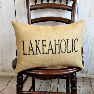 May include: A burlap pillow with the text "LAKEAHOLIC" printed in black on a brown wooden chair.