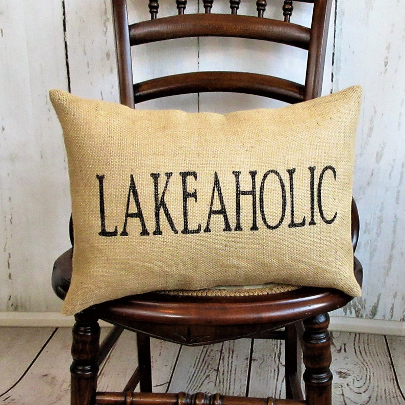 May include: A burlap pillow with the text "LAKEAHOLIC" printed in black on a brown wooden chair.