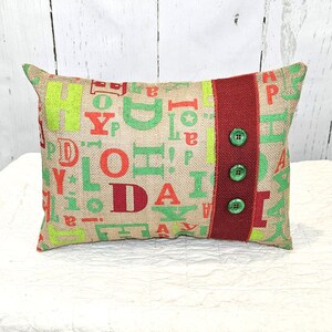 May include: Rectangular burlap pillow with green and red letters spelling "JOY" and "DA". A vertical red stripe with three green buttons is on the right side. The pillow is a decorative item.