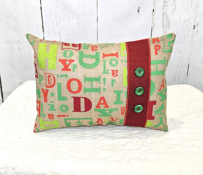 May include: Rectangular burlap pillow with green and red letters spelling "JOY" and "DA". A vertical red stripe with three green buttons is on the right side. The pillow is a decorative item.