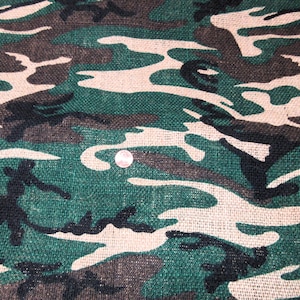 May include: Green, brown, and black camouflage fabric with a burlap texture. The fabric has a repeating pattern of irregular shapes.