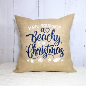 May include: A tan burlap throw pillow with the text "Have Yourself a Beachy Little Christmas" in blue lettering and white seashells and stars.