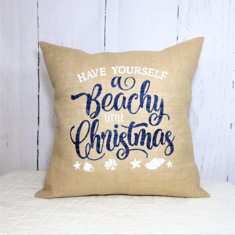 May include: A tan burlap throw pillow with the text "Have Yourself a Beachy Little Christmas" in blue lettering and white seashells and stars.