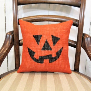 May include: Orange burlap cushion with a black jack-o'-lantern face design. The cushion is sitting on a striped chair.