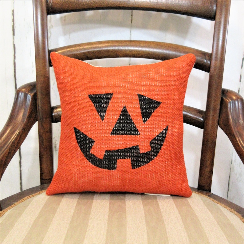May include: Orange burlap cushion with a black jack-o'-lantern face design. The cushion is sitting on a striped chair.