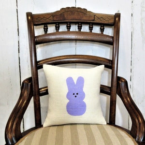 May include: A white pillow with a purple bunny rabbit design. The pillow is sitting on a wooden chair with a striped seat.