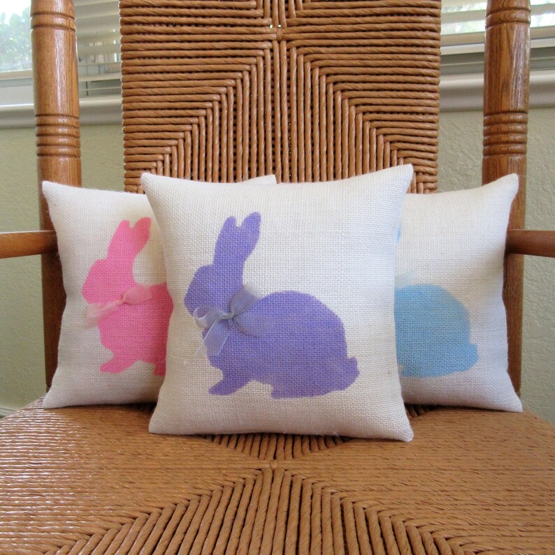 May include: Three decorative throw pillows with bunny silhouettes. The pillows are made of a natural linen fabric and feature a pink, purple, and blue bunny design.