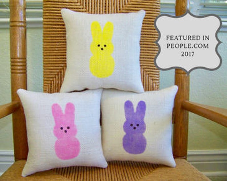 May include: Three decorative throw pillows with a bunny-shaped design. The pillows are made of a light-colored fabric and feature a yellow, pink, and purple bunny design.