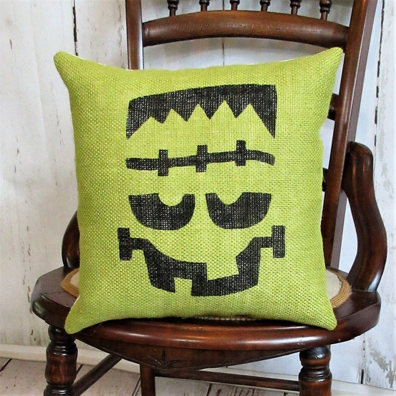 May include: Green burlap pillow with a black Frankenstein face design. The pillow is sitting on a wooden chair.