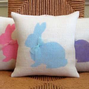May include: Three white cushion covers with bunny silhouettes. The bunny on the left is pink, the middle bunny is blue, and the bunny on the right is purple.