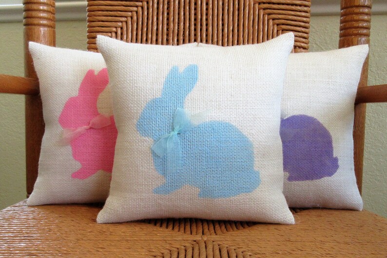 May include: Three white cushion covers with bunny silhouettes. The bunny on the left is pink, the middle bunny is blue, and the bunny on the right is purple.