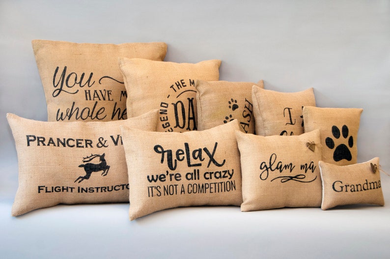 May include: A collection of burlap throw pillows with various sayings and designs, including "You have my whole heart", "The Legend - Dad", "Prancer & Vixen - Flight Instructor", "Relax we're all crazy it's not a competition", "Glam Ma", and "Grandma".