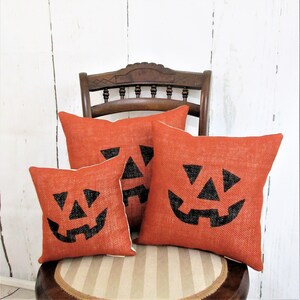 May include: Three orange burlap throw pillows with black jack-o'-lantern faces on a wooden chair. The pillows are arranged in a pyramid shape, with the largest pillow at the bottom and the smallest pillow at the top.