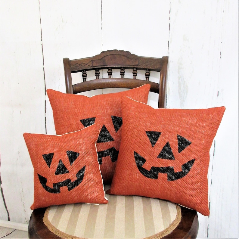 May include: Three orange burlap throw pillows with black jack-o'-lantern faces on a wooden chair. The pillows are arranged in a pyramid shape, with the largest pillow at the bottom and the smallest pillow at the top.