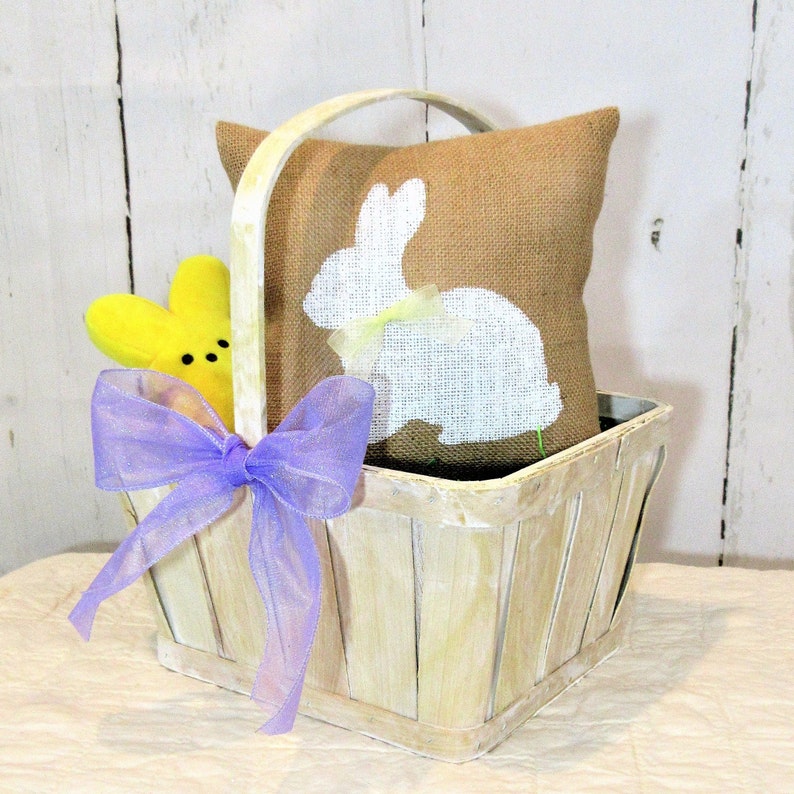 Puede incluir: Una cesta de madera blanqueada con una cinta morada y una almohada de arpillera con una silueta de conejo blanco. Tambi&eacute;n hay un juguete de pollito amarillo en la cesta.