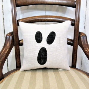 May include: A white cushion with a black ghost face design. The cushion is made from a burlap-like fabric and has a simple, minimalist design.