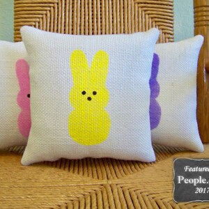May include: Three decorative throw pillows with a woven texture. The pillows are white with a pink, yellow, and purple bunny design. The pillows are sitting on a wicker chair. The text "Featured on People.com 2017" is on a black chalkboard.