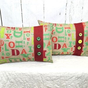 May include: Two rectangular burlap pillows with a festive design. The pillows feature a mix of green and red letters spelling out "JOY" and "DAY". A red stripe with green and yellow buttons runs down the center of each pillow. The pillows are on a white surface.
