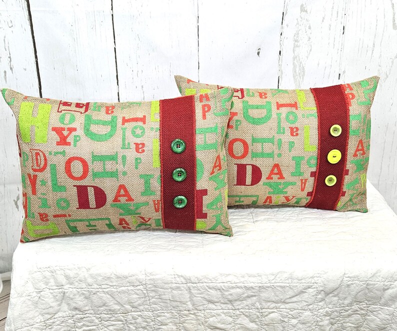 May include: Two rectangular burlap pillows with a festive design. The pillows feature a mix of green and red letters spelling out "JOY" and "DAY". A red stripe with green and yellow buttons runs down the center of each pillow. The pillows are on a white surface.