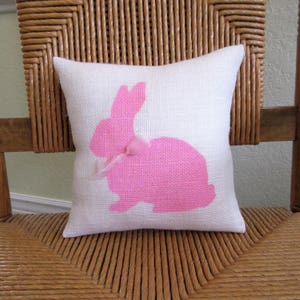 May include: A white cushion with a pink bunny silhouette. The bunny has a pink ribbon around its neck.