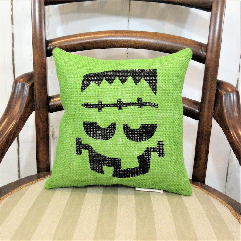 May include: Green burlap cushion with a black Frankenstein face design. The cushion is sitting on a striped chair.