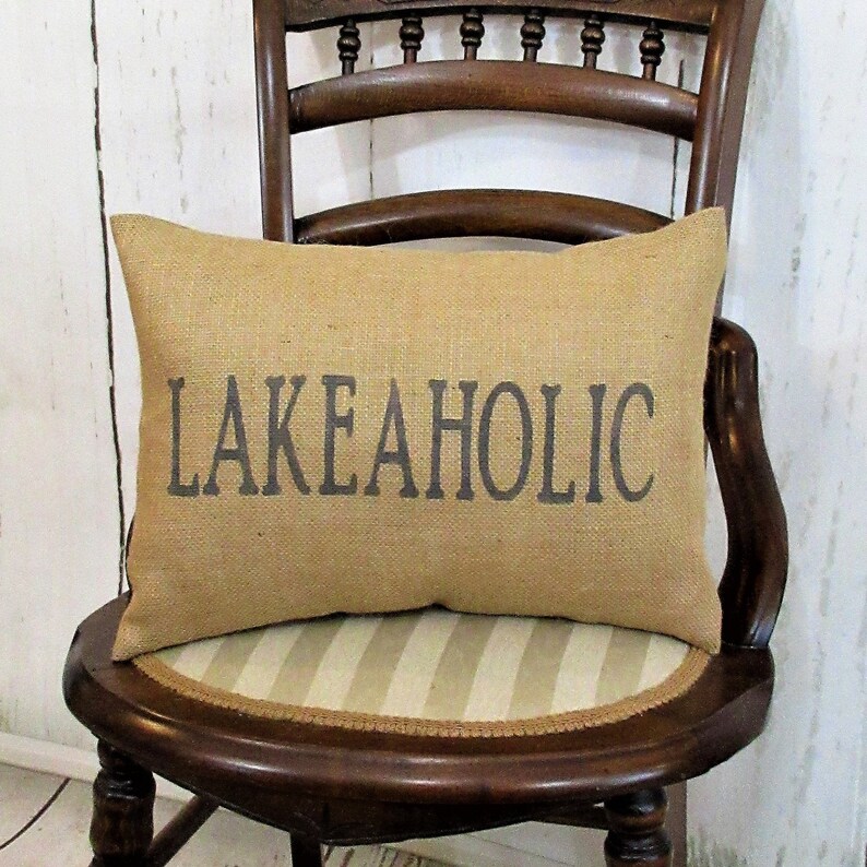 May include: A brown burlap pillow with the text "LAKEAHOLIC" printed in gray on the front. The pillow is sitting on a wooden chair with a striped seat.
