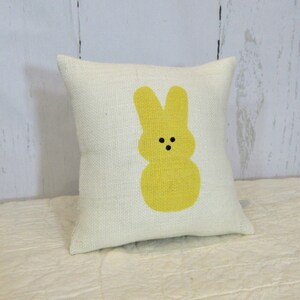 May include: A white pillow with a yellow bunny rabbit design. The bunny has black eyes and a simple, rounded shape.