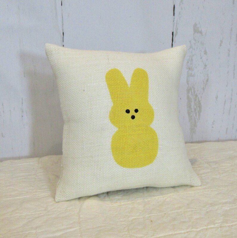 May include: A white pillow with a yellow bunny rabbit design. The bunny has black eyes and a simple, rounded shape.