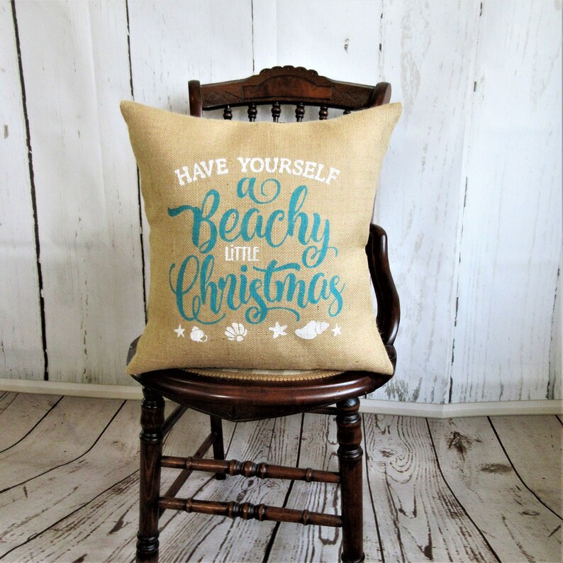May include: A burlap pillow with the text "Have Yourself a Beachy Little Christmas" in teal blue lettering. The pillow is decorated with white stars and seashells.