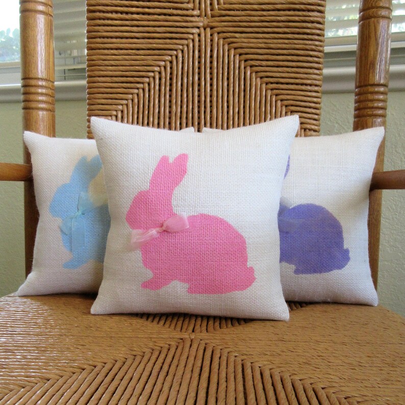 May include: Three decorative throw pillows with white fabric and bunny silhouettes. The bunnies are pink, blue, and purple. The pillows are sitting on a wicker chair.