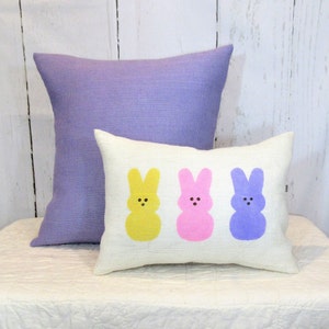 May include: Three decorative throw pillows with a white background and three Easter bunny silhouettes. The bunnies are yellow, pink, and purple.
