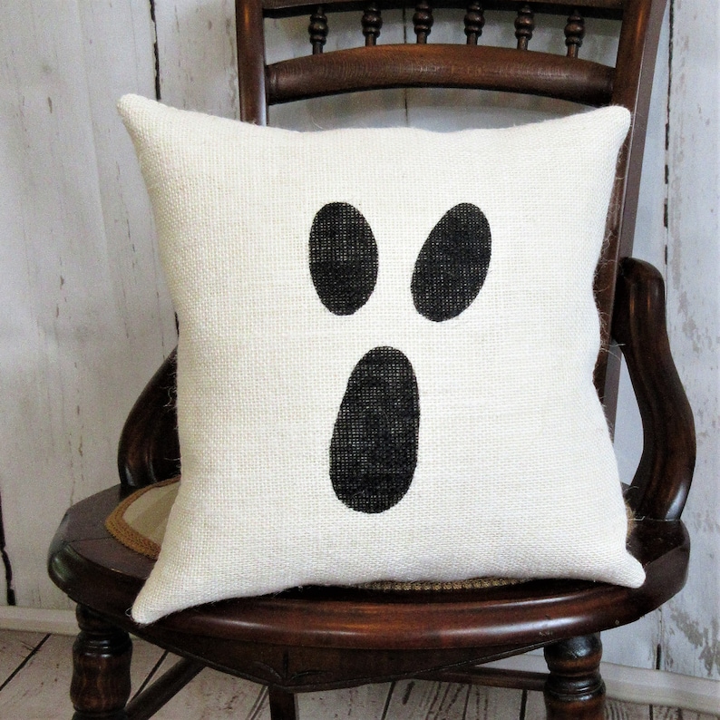 May include: A white pillow with a black ghost face design. The pillow is made of burlap and has a simple, rustic look.