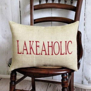 May include: A beige burlap pillow with the word "LAKEAHOLIC" in red lettering. The pillow is sitting on a wooden chair.