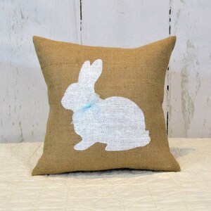 Puede incluir: Una almohada de arpillera con una silueta blanca de un conejo con un lazo azul. La almohada es perfecta para la decoraci&oacute;n de primavera.