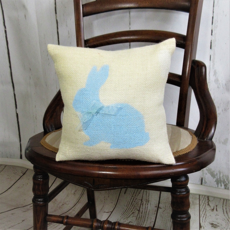 May include: A light blue bunny silhouette on a cream-coloured burlap cushion. The bunny has a light blue bow on its back. The cushion is sitting on a wooden chair.