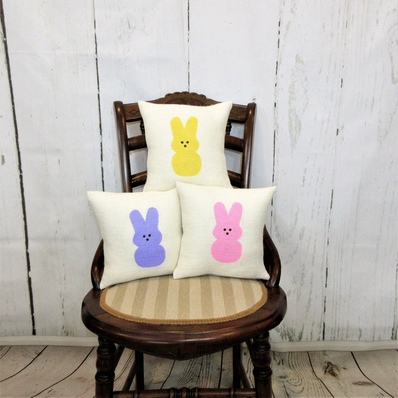 May include: Three decorative throw pillows with a white background and a pastel colored bunny rabbit design. The pillows are arranged on a wooden chair with a striped seat.