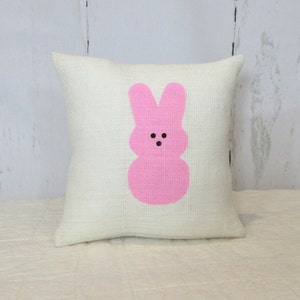 May include: A white pillow with a pink bunny rabbit design. The bunny has a simple face with two black eyes and a small mouth.