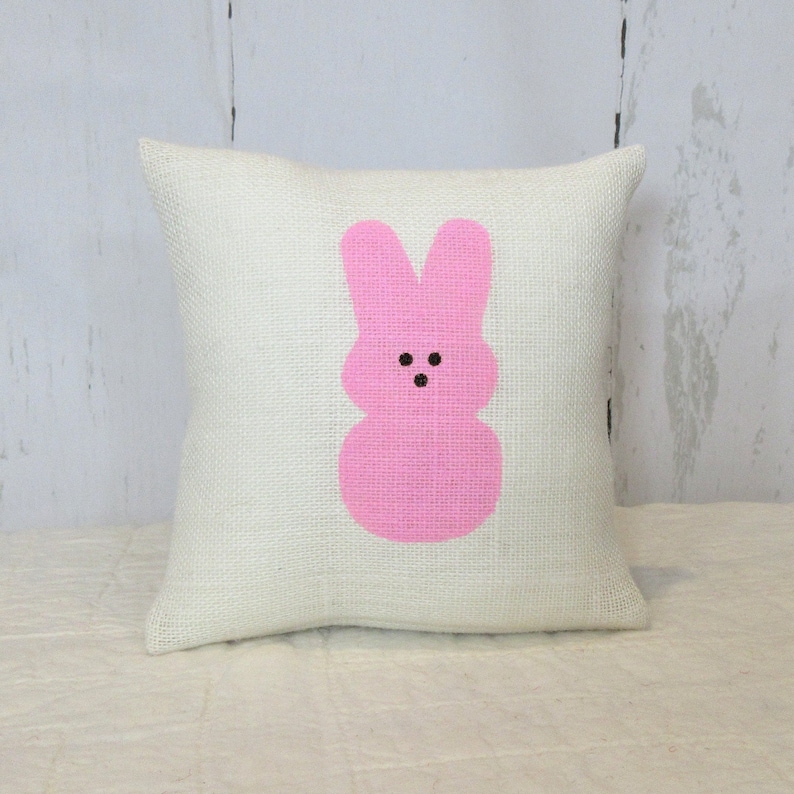 May include: A white pillow with a pink bunny rabbit design. The bunny has a simple face with two black eyes and a small mouth.