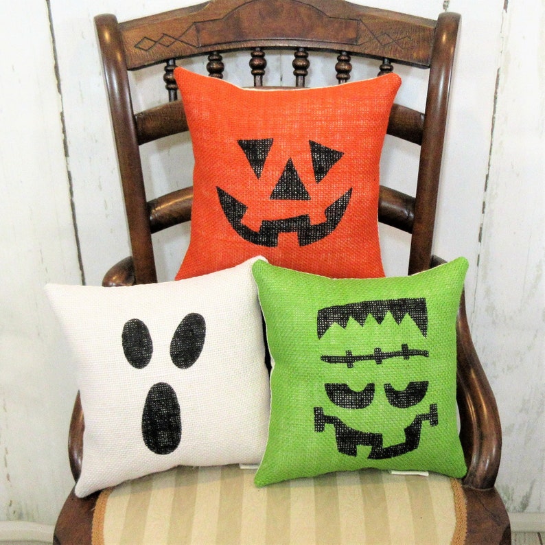 May include: Three decorative throw pillows with Halloween-themed faces. The pillows are made of burlap and feature a pumpkin, a ghost, and a Frankenstein monster. The pillows are arranged on a wooden chair with a striped seat.