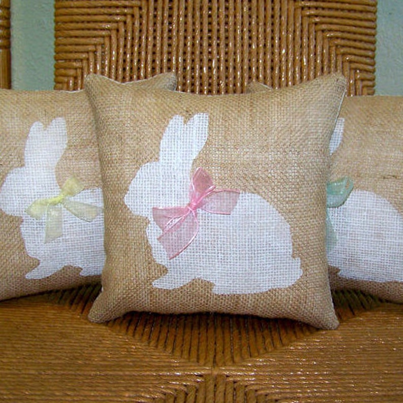 Puede incluir: Tres fundas de coj&iacute;n de arpillera con siluetas de conejos blancas y lazos de cinta de color pastel. Los cojines est&aacute;n dispuestos en una silla de mimbre tejido.