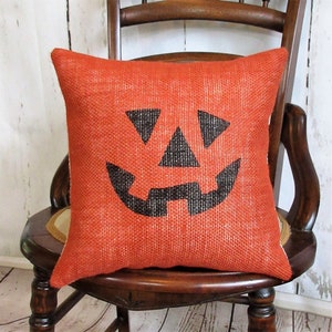 May include: Orange burlap pillow with a black jack-o'-lantern face design. The pillow is sitting on a wooden chair.
