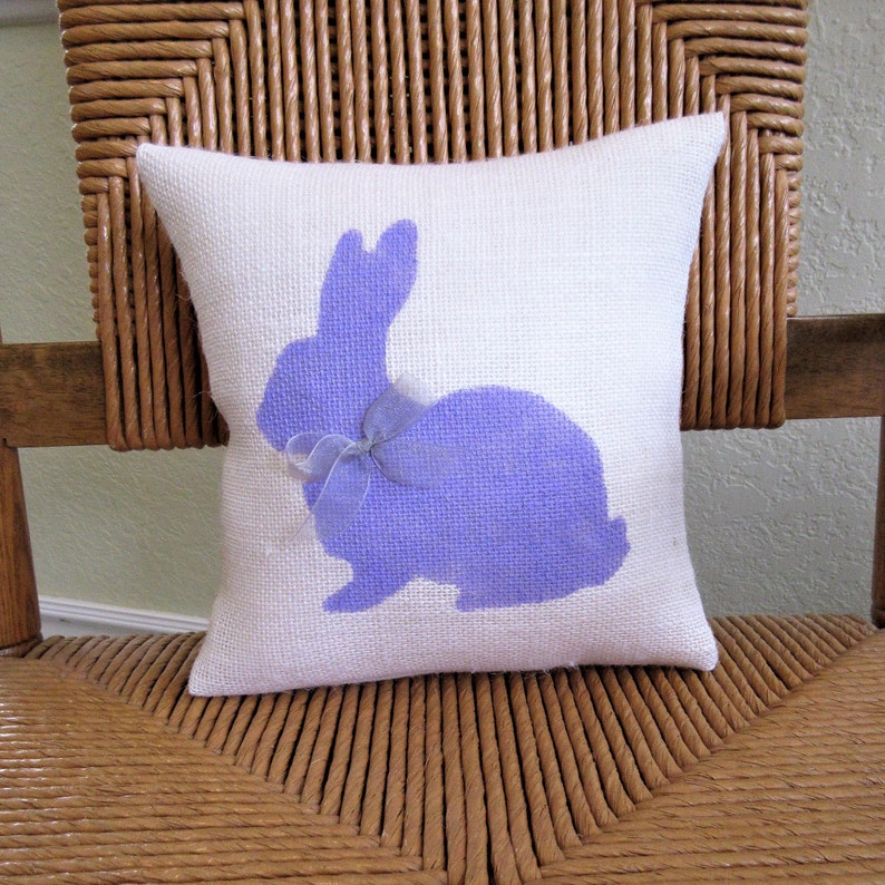 May include: A white pillow with a purple bunny silhouette and a purple bow. The pillow is made of burlap and has a rustic look.