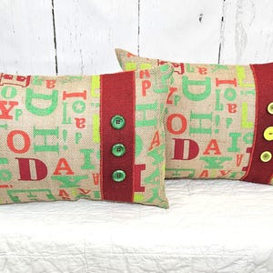 May include: Two rectangular burlap pillows with a festive design. The pillows feature a repeating pattern of green and red letters. A red stripe with green buttons adorns one pillow, while the other has yellow buttons.