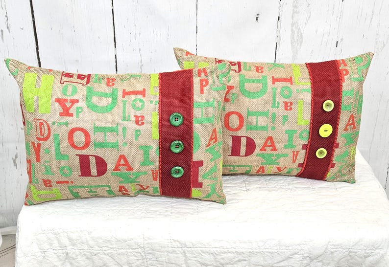 May include: Two rectangular burlap pillows with a festive design. The pillows feature a repeating pattern of green and red letters. A red stripe with green buttons adorns one pillow, while the other has yellow buttons.